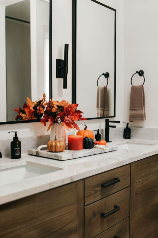 Fall-themed bathroom vanity decorated with a white vase of orange autumn leaves, assorted mini pumpkins, pillar candles, and hand soap bottles arranged on a marble tray. The warm wood cabinetry, matte black fixtures, and neutral towels create a cozy seasonal vibe. 