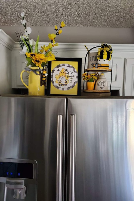 Bee-themed decor above a stainless steel refrigerator featuring a yellow pitcher with faux flowers, a framed “Bee Kind” sign, and a two-tiered tray with honey jars, floral accents, and a gnome wearing a bee outfit. 