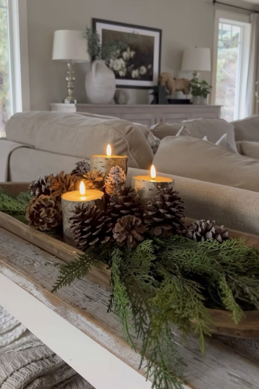 Cozy winter dough bowl centerpiece styled with lit birch-wrapped candles, pinecones, and fresh cedar greenery, displayed on a distressed white console table in a warm neutral living room.