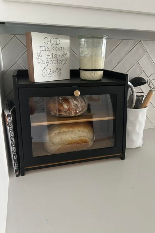 Modern farmhouse bread station featuring a black bread display box with loaves of artisan bread, a wooden faith sign with Psalm 28:7, sourdough starter in a Weck jar, and a ceramic utensil holder on a clean white counter and chevron tile backsplash