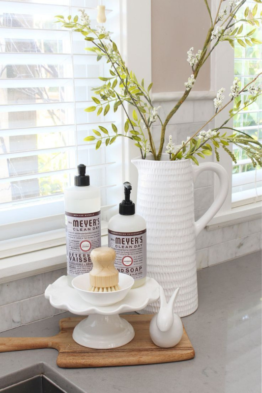 Bright kitchen sink decor setup with Mrs. Meyer’s hand and dish soap, wooden scrub brush, fluted pitcher with tall green stems, all arranged on a wood cutting board and white pedestal tray by a sunlit window with shutters
