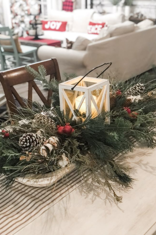 Festive Christmas dough bowl centerpiece featuring a white lantern with a glowing candle, surrounded by snowy pinecones, red berries, birch logs, and lush evergreen branches, styled on a striped table runner in a cozy holiday-themed living and dining space.