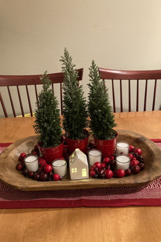Festive Christmas dough bowl centerpiece on a wooden dining table, featuring mini potted evergreen trees in red containers, white votive candles, a ceramic house lantern, and scattered red berries arranged over a red woven table runner.