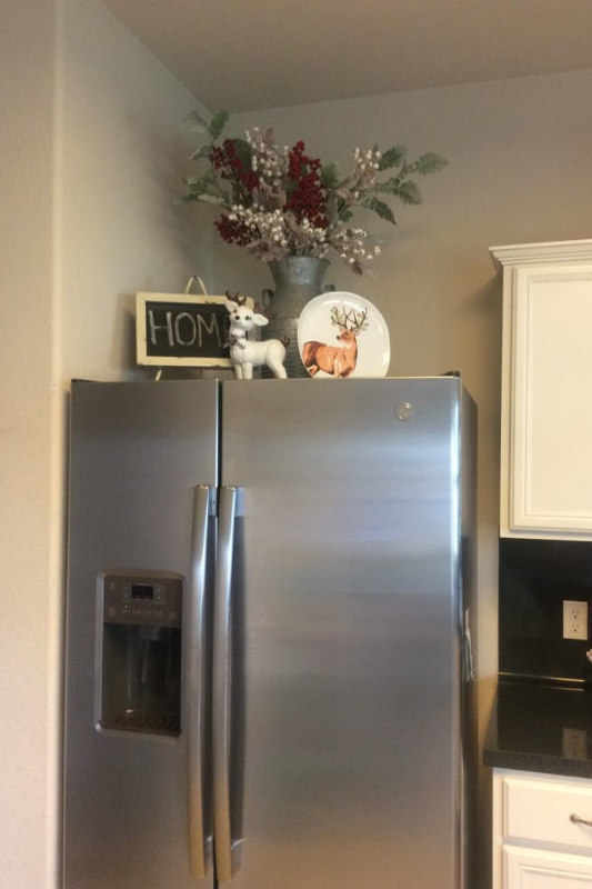 Festive kitchen with stainless steel fridge topped with holiday decor including a faux reindeer, “HOME” chalkboard sign, floral arrangement, and decorative reindeer plate 