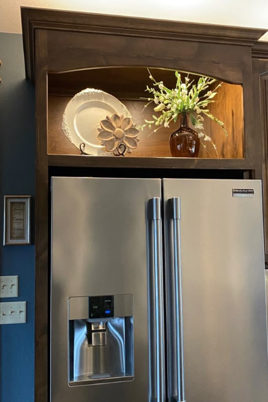 Built-in wooden display shelf above stainless steel fridge featuring a decorative plate, metal flower, and floral arrangement for an elegant kitchen look