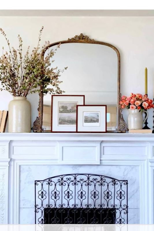 Elegant white fireplace mantel styled with a vintage gold-framed mirror, blush floral arrangements, tall gold taper candles, ceramic vases, and framed art prints. This timeless setup blends French country charm and transitional decor, ideal for spring mantle styling, traditional home inspiration, and curated vintage interior looks.