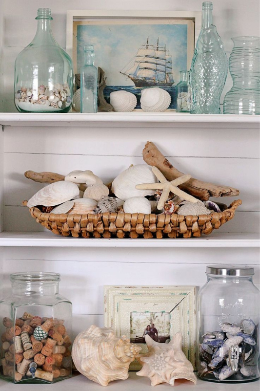 Beach-inspired bookshelf styling with glass bottles, driftwood, woven basket of seashells, nautical ship painting, and vintage coastal accessories 