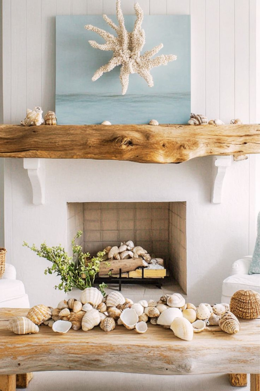 Bright coastal fireplace featuring a raw driftwood mantel adorned with scattered seashells, set against white shiplap walls. A striking coral-inspired sculpture mounted on an ocean-blue canvas creates a serene focal point, while the hearth and coffee table overflow with large natural shells and green accents—ideal for beach house interiors, nautical living room inspiration, and summer fireplace decor ideas.