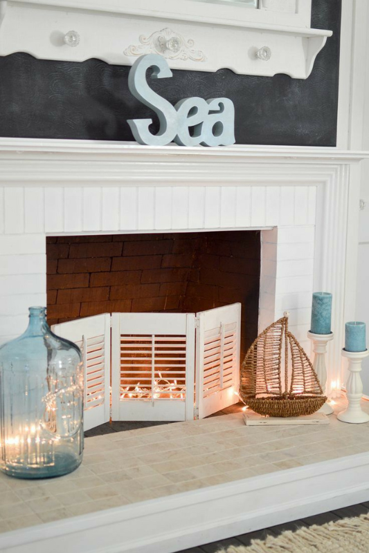 Light and breezy coastal fireplace decor featuring a white brick surround, soft blue "Sea" sign, large glass jug with twinkle lights, and a wicker sailboat. Blue pillar candles on white stands and a shutter-style fireplace screen complete the seaside theme—perfect for beach cottage inspiration, nautical living room ideas, and relaxed summer mantel styling.