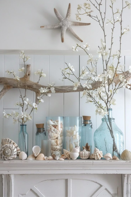Serene coastal-inspired fireplace mantel decorated with pastel seashells, clear blue glass vases, and blooming white branches arranged around natural driftwood. A woven starfish wall accent completes the breezy seaside aesthetic—perfect for beach house decor, summer mantel styling, and relaxed nautical interior design ideas.