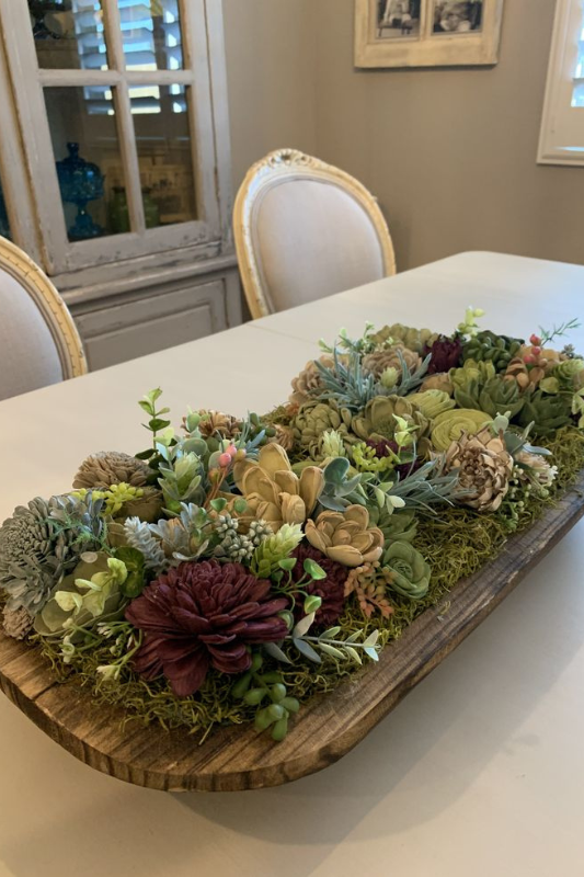 Vibrant spring dough bowl centerpiece arranged with a variety of faux flowers in shades of burgundy, green, beige, and blue, nestled in moss on a rustic wooden bowl placed on an elegant white dining table in a French country-style dining room.