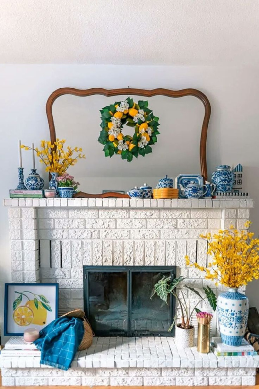 Charming white brick fireplace styled with a vibrant lemon wreath, curved vintage mirror, and bold blue-and-white chinoiserie vases. Accents include yellow florals, playful lemon artwork, and layered spring books, making it a cheerful seasonal mantle perfect for bright farmhouse interiors, citrus-inspired decor, and eclectic living room styling.