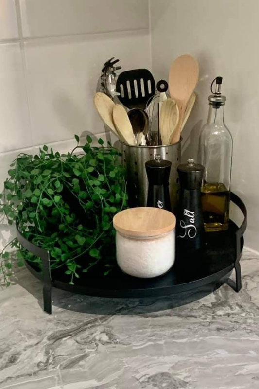 Functional and cozy kitchen corner styling with a round black tray holding a faux trailing plant, olive oil bottle, salt and pepper shakers, a white canister with wooden lid, and a metal utensil holder, all placed on a marbled gray countertop