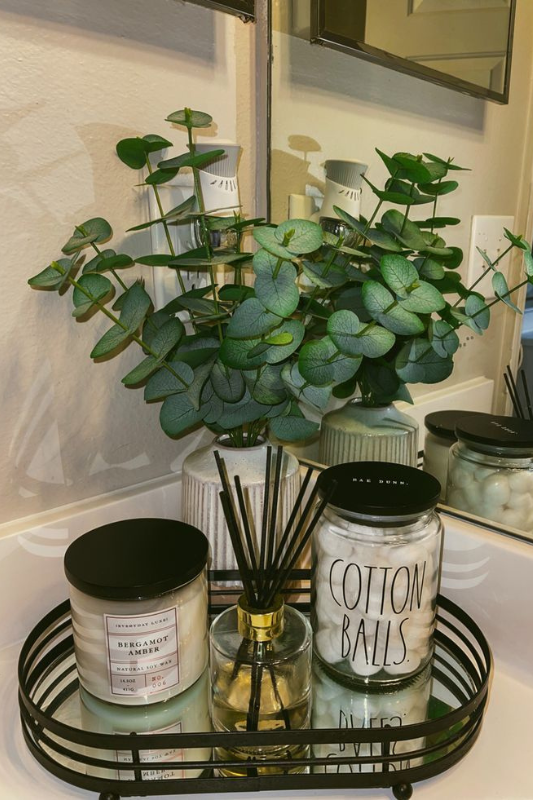 Cozy bathroom countertop decor featuring a round black tray with a eucalyptus arrangement in a ribbed vase, a Rae Dunn cotton ball jar, a reed diffuser, and a Bergamot Amber candle—styled in front of a mirror for a warm and inviting display. 