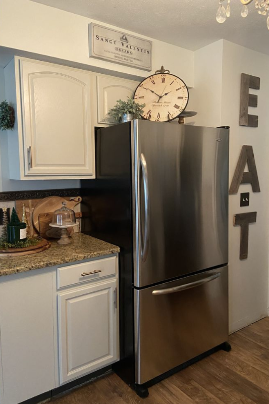 Farmhouse-style kitchen with stainless steel fridge topped with a large vintage clock, greenery, and a rustic bread sign for cozy wall decor