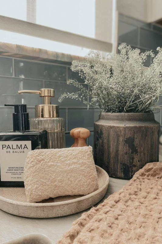 Earthy bathroom decor featuring a ceramic dish with a textured washcloth, Palma hand sanitizer, glass soap dispenser with gold pump, wooden massage brush, and a rustic vase filled with soft green dried florals. The setup is complemented by a waffle-knit towel, creating a warm and natural aesthetic. 