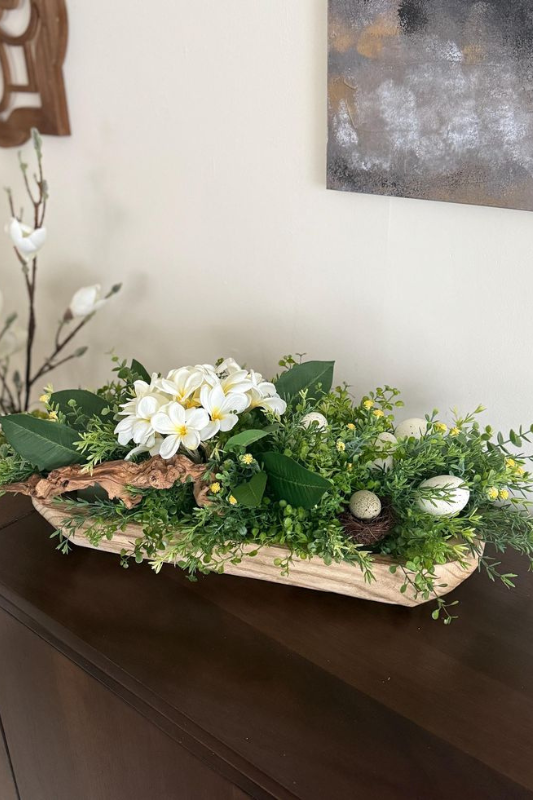 Fresh Easter dough bowl centerpiece featuring white primrose flowers, lush green foliage, decorative eggs, and a small bird nest, arranged in a light wooden bowl and displayed on a dark wood console table in a spring-themed home.