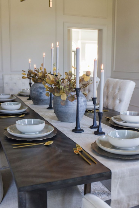 Elegant fall dining table centerpiece featuring tall black taper candle holders, vintage-style ceramic vases with golden eucalyptus stems, and a neutral linen runner styled with gold flatware and layered place settings. 