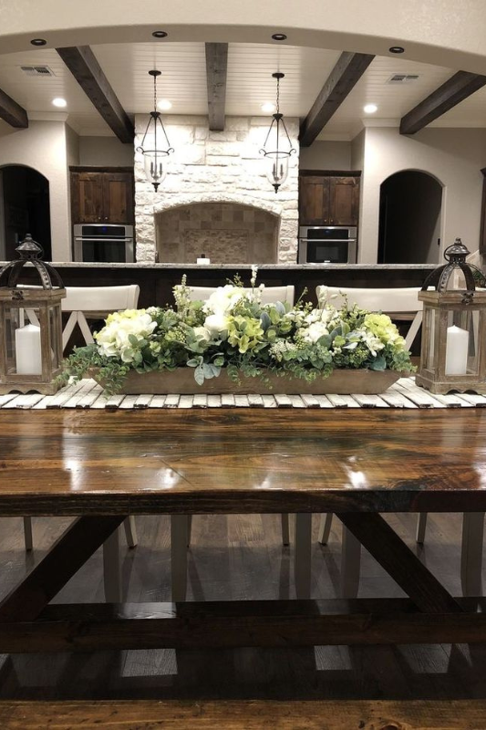 Elegant dough bowl centerpiece filled with white and green faux flowers and eucalyptus stems, flanked by rustic lanterns with pillar candles, styled on a wood slat runner atop a dark farmhouse dining table in a cozy open-concept kitchen and dining space.
