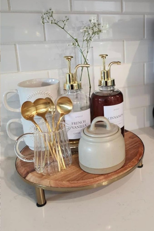 Elegant kitchen sink styling with a marble tray holding a burning Diptyque candle, stylish white soap dispensers with gold pumps, and a sleek gooseneck faucet on a speckled granite countertop with ambient lighting