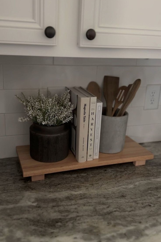 Minimalist elevated kitchen counter decor featuring a wooden riser with neutral-toned cookbooks, a rustic black planter filled with faux white florals, and a cement utensil holder with wooden kitchen tools against a white subway tile backsplash