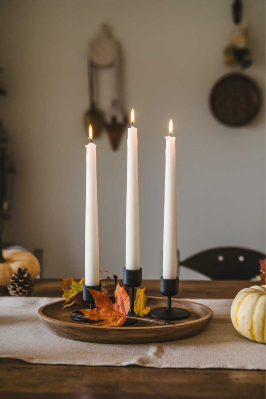 Minimalist fall dining table centerpiece with three black metal candleholders, white taper candles, and decorative autumn leaves on a wooden tray for a cozy seasonal tablescape. 