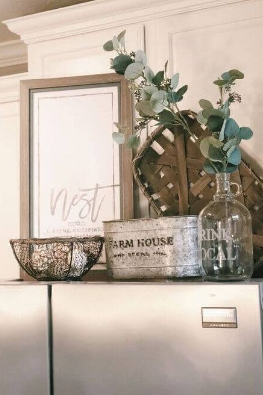 Farmhouse-style vignette above a fridge featuring a framed “Nest” sign, tobacco basket, greenery in a glass bottle, a galvanized metal container labeled “Farm House,” and a rustic wire bowl. 