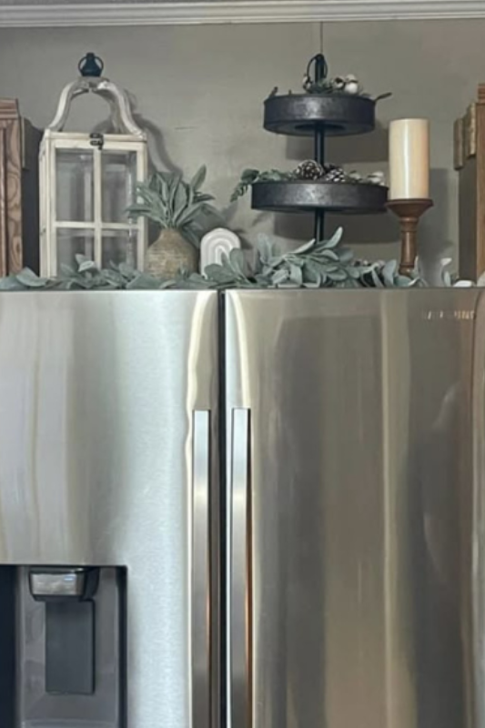 Rustic farmhouse above-fridge decor featuring a black metal tiered tray, white candle on a wooden holder, small vintage window frame, greenery garland, and neutral accent pieces. 