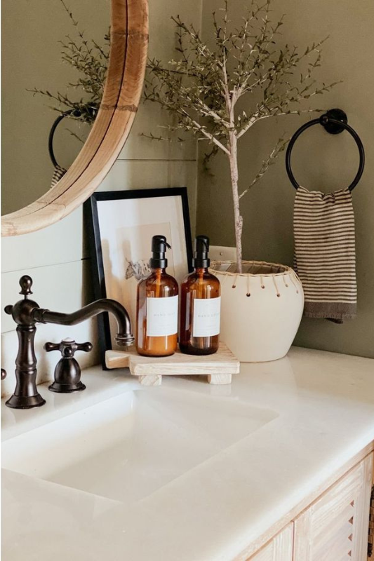 Farmhouse-style bathroom vanity with a white countertop, bronze faucet, and wooden-framed mirror, styled with amber soap dispensers on a rustic riser, a potted faux tree, striped hand towel, and framed art for a warm, earthy, and timeless look. 