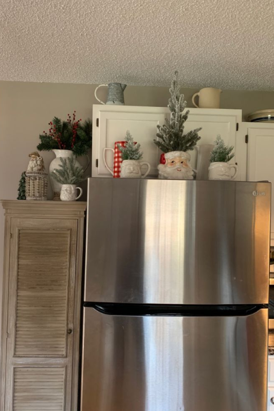 Farmhouse-style Christmas kitchen decor featuring a stainless steel fridge topped with festive mugs, mini flocked trees, greenery, and a Santa cookie jar on white cabinets. 