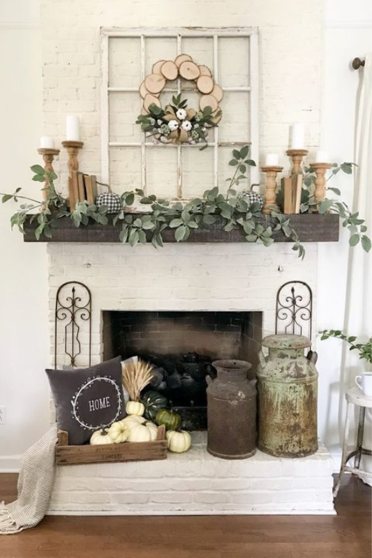Charming farmhouse fireplace styled with a rustic wooden mantel, draped eucalyptus garland, and vintage wood candlesticks. Accents include stacked books, gingham pumpkins, an antique window frame with a wood slice wreath, and vintage milk jugs. A crate filled with pumpkins, wheat, and a "Home" pillow adds cozy fall charm—perfect for rustic seasonal styling, farmhouse living room inspiration, and autumn mantel decor ideas.