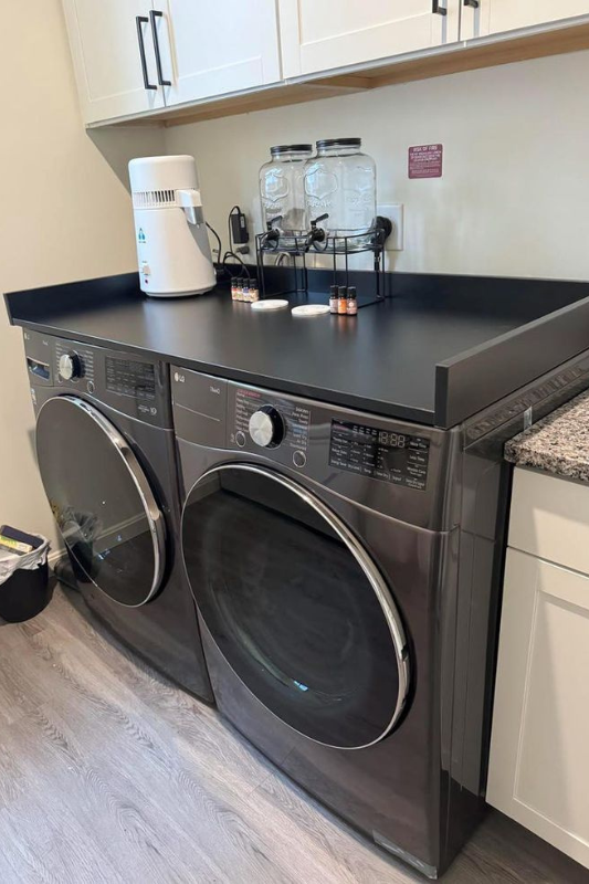 Charming farmhouse laundry room with botanical wallpaper, open wood shelving, indoor plants, and front-load washer and dryer, creating a cozy and inviting vibe. 