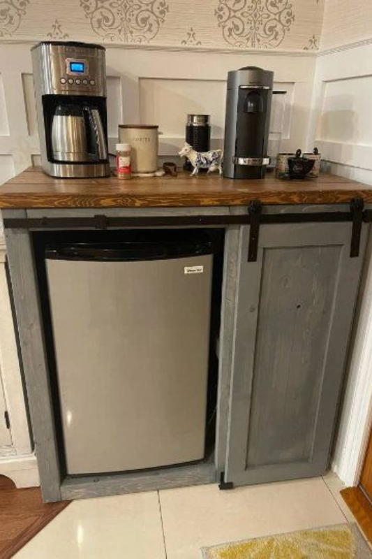 Rustic farmhouse coffee bar featuring a mini fridge enclosed in a gray sliding barn door cabinet, topped with a wood counter holding a coffee maker, Keurig, and mugs. 