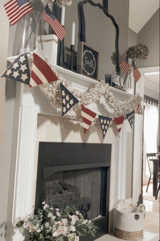 Festive Fourth of July fireplace mantel decorated with patriotic flag bunting, mini American flags, neutral ruffle garlands, and vintage-inspired floral arrangements. A black-framed mirror and farmhouse-style "Faith" sign anchor the look, blending Americana charm with cozy rustic touches—ideal for Independence Day decor, summer holiday inspiration, and farmhouse living room styling.