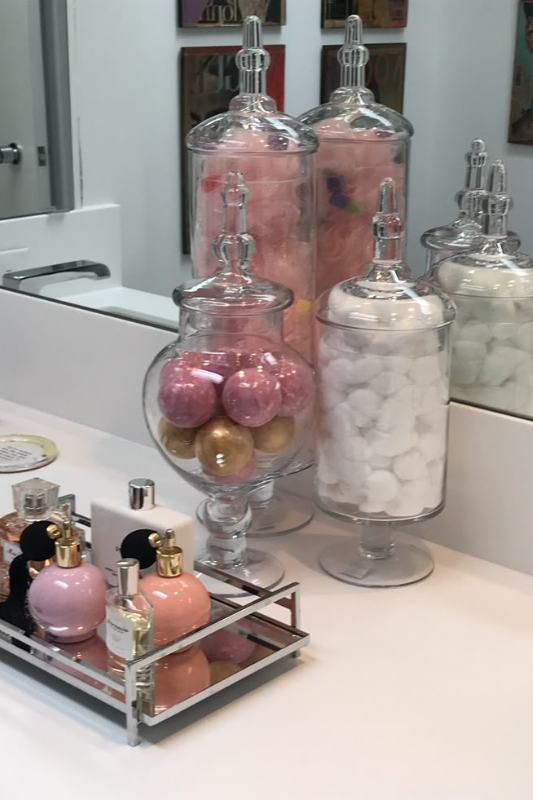 Glamorous bathroom counter decorated with tall glass apothecary jars filled with cotton balls, bath bombs, and decorative items, alongside a mirrored tray displaying pink and gold perfume bottles. The setup adds a luxurious, feminine touch with a sleek and polished finish. 