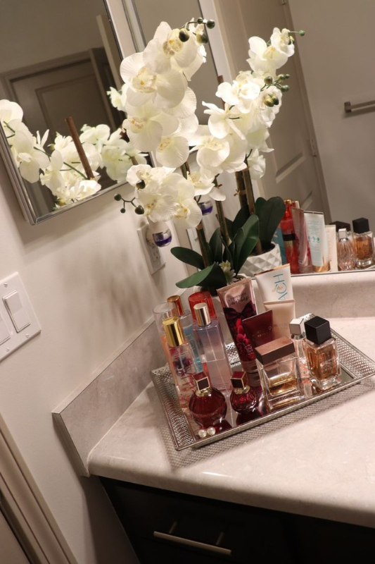 Stylish bathroom countertop decor featuring a silver mirrored tray filled with assorted perfumes, body sprays, and lotions, alongside a tall arrangement of white orchids in a textured pot. The setup creates a glamorous, feminine display that’s both organized and luxurious. 