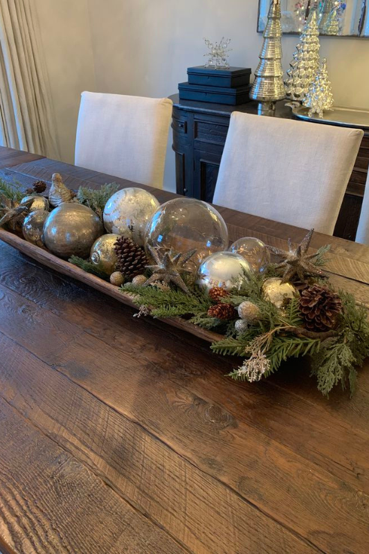 Sophisticated Christmas dough bowl centerpiece on a rustic wood dining table, filled with gold and clear ornaments, pinecones, glittery stars, and evergreen branches, creating an elegant and festive holiday display.