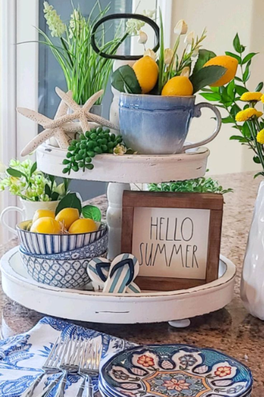 Coastal summer tiered tray decor with faux lemons, blue mugs and bowls, starfish accents, and a “Hello Summer” sign styled on a kitchen countertop with patterned blue dishes and napkins 