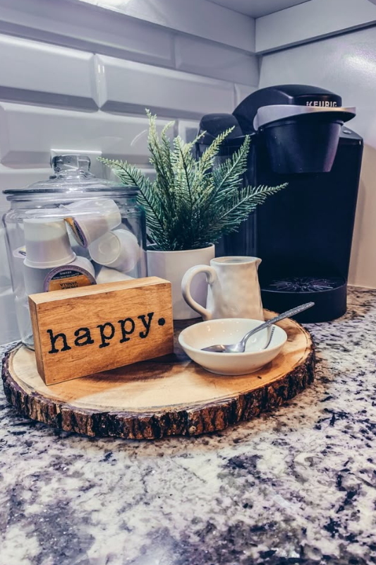 Rustic coffee corner featuring a Keurig coffee maker, glass jar filled with pods, mini evergreen plant, ceramic creamer, and a wood slice tray with a “happy” sign for a cozy and inviting coffee setup on granite countertops