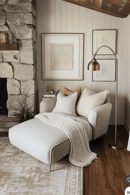 Living room corner with cream lounge chair, knit throw, stone fireplace, shiplap walls, brass arc floor lamp, and framed botanical art
