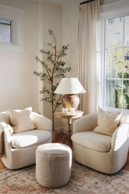 Living room corner with two boucle swivel chairs, knit ottoman, round wood table, ceramic lamp, and faux olive tree near sunlit window