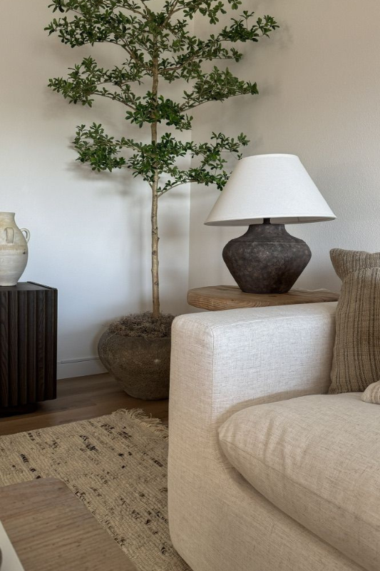 Neutral living room corner with tall faux tree, round ceramic lamp on wood side table, textured rug, and soft beige sofa