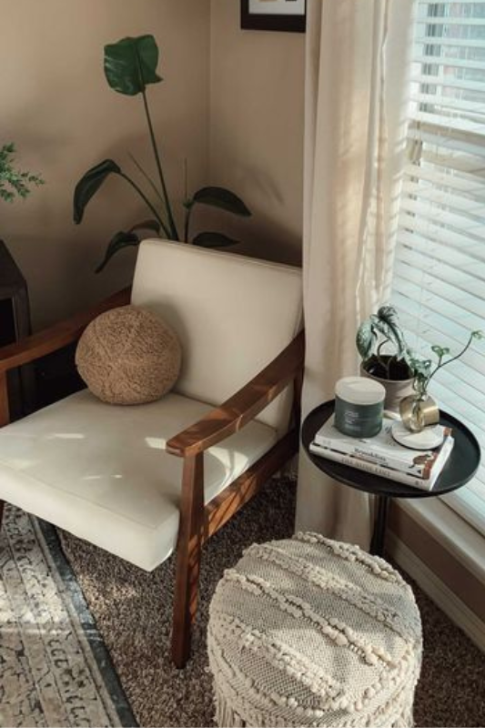 Cozy living room corner with wood armchair, round pouf, black side table, candles, plants, and soft neutral curtains with natural light 