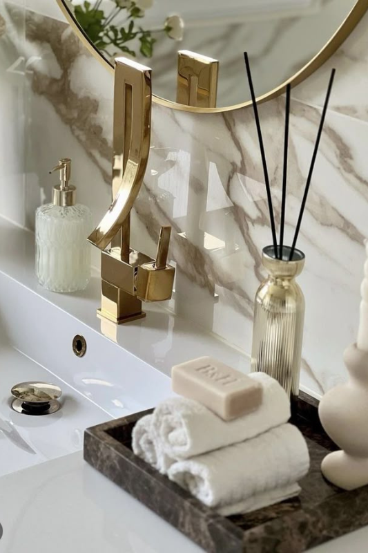 Elegant bathroom vanity styled with a striking gold faucet, marble backsplash, and a dark marble tray holding rolled towels, a bar of soap, a sculptural candle, and a reed diffuser. A vintage glass soap dispenser and floral mirror reflection enhance the space’s luxurious, spa-like feel. 
