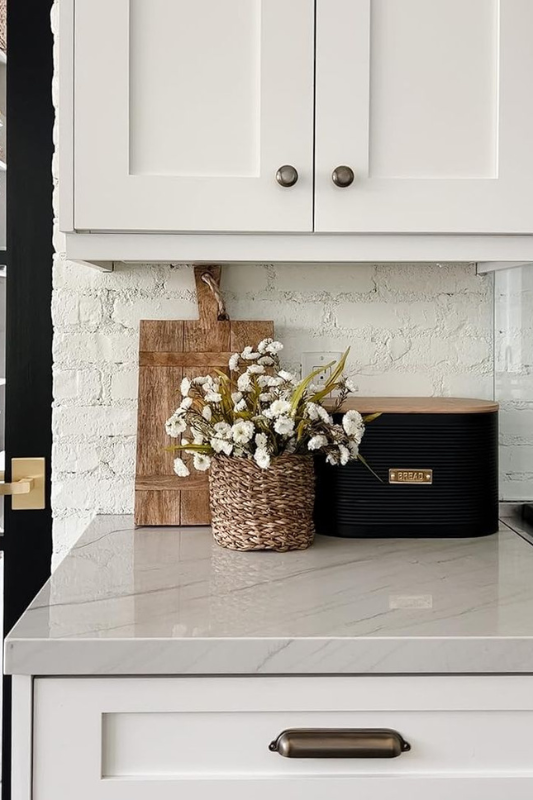 Minimalist modern kitchen counter decor featuring a black ribbed bread box with wood lid, a woven basket filled with white faux florals, and a rustic wood cutting board leaning against a painted white brick backsplash under white shaker cabinets