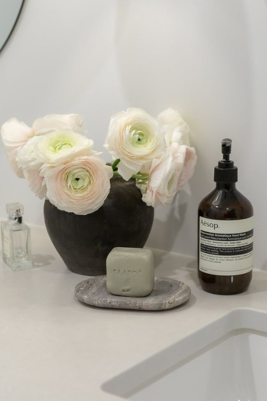 Minimalist bathroom countertop featuring a black vase of pale blush ranunculus flowers, a green square bar of soap on a natural stone dish, a glass perfume bottle, and a brown Aesop hand wash bottle. The soft, neutral tones and curated arrangement create a serene and elegant look. 