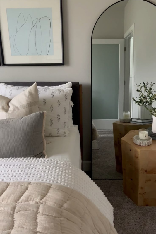 Minimalist bedroom corner featuring a full-length arched mirror, soft neutral bedding with textured throw pillows, modern wooden side table with candles and greenery, and abstract wall art for a calm and stylish space. 
