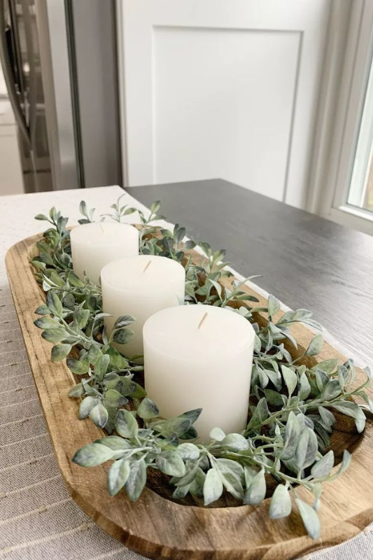 Minimalist dough bowl centerpiece on a modern farmhouse dining table, featuring three white pillar candles surrounded by soft green faux leaves, placed on a neutral striped table runner near a bright kitchen window.