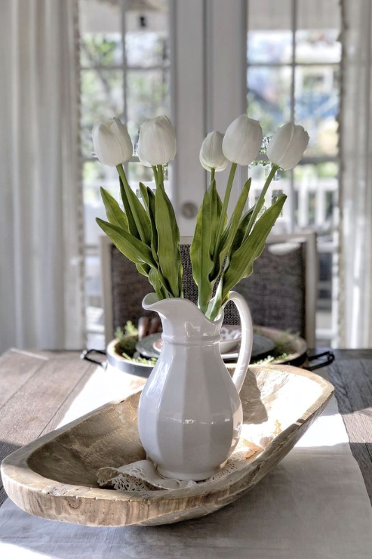 Simple and elegant dough bowl centerpiece featuring a white ceramic pitcher holding fresh white tulips, placed on a neutral table runner in a bright farmhouse dining room with natural light streaming through French doors.