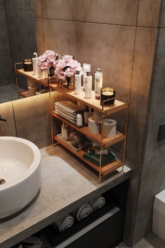 Modern bathroom countertop featuring a gold-framed tiered shelf with skincare products, rolled towels, candles, and pink roses in a glass vase. The concrete countertop and wall tiles add a sleek, contemporary touch to the warm and organized display. 